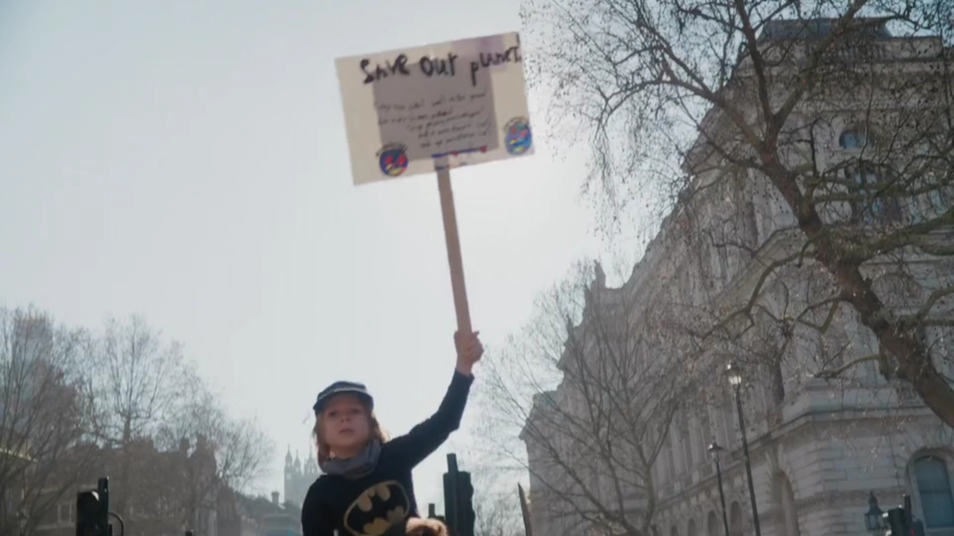 A young climate protestor wearing a batman jumper and holding a placard saying Save Our Planet