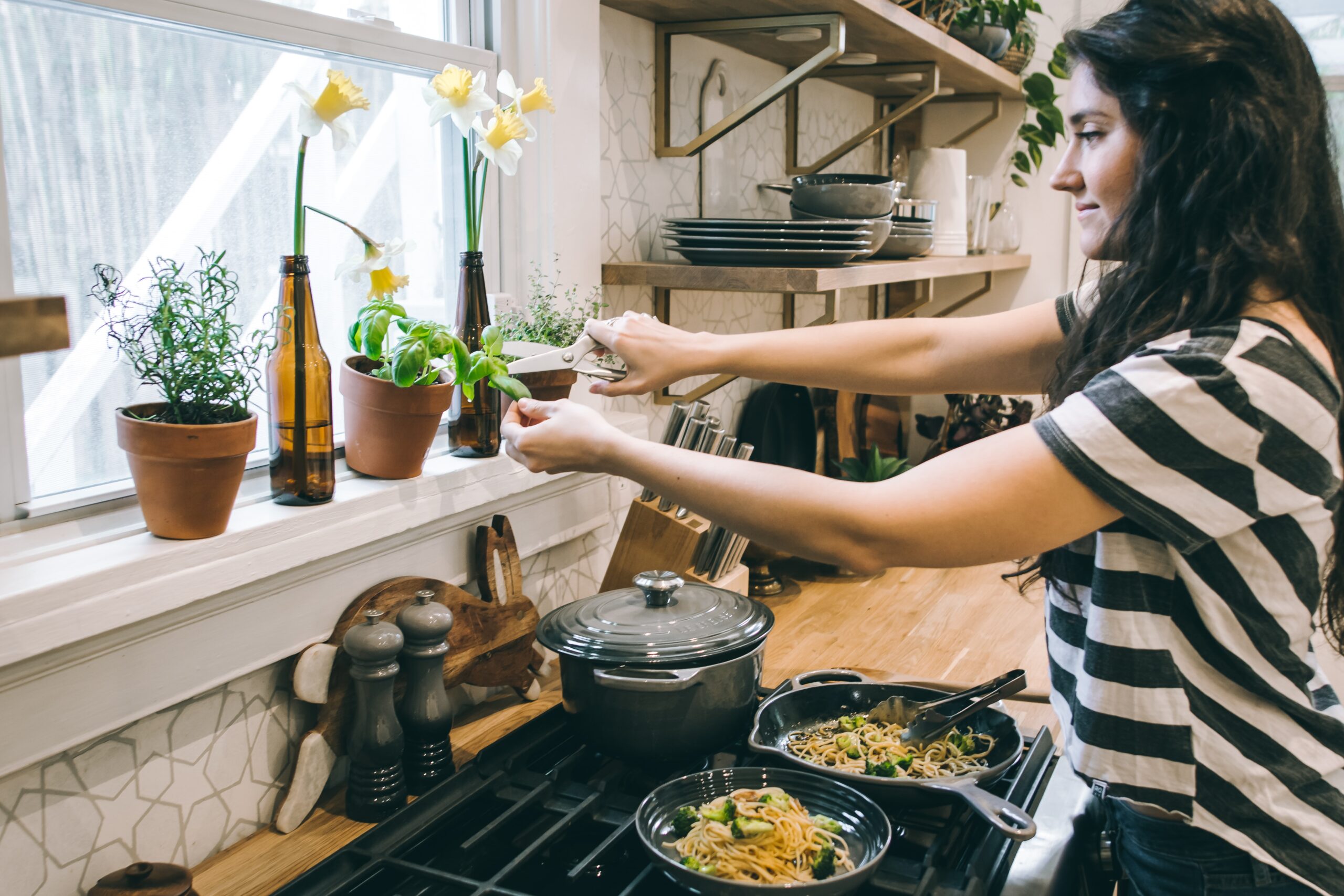 Photo of a woman cooking, cutting a basil leaf from a plant, by Tina Dawson on Unsplash